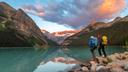Zwei Wanderer mit großen Rucksäcken stehen am felsigen Ufer und blicken auf einen türkisfarbenen Gebirgssee. Steile, bewaldete Berge rahmen das Wasser ein. Die fernen Gipfel leuchten im goldenen Abendlicht. Die ruhige Oberfläche spiegelt die Landschaft und den rosa-orangen Himmel klar wider.