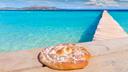 Ein mallorquinischer Schmalzkuchen auf einer Mauer am Meer. Das Wasser ist hellblau und die Sonne scheint.