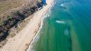 Luftaufnahme vom Vaya Beach mit weitem, hellem Sandstrand, türkisfarbenem Meer und sanften Wellen, umgeben von naturbelassener Küstenlandschaft.