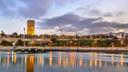 Der beleuchtete Hassan-Turm in Rabat erhebt sich bei Abenddämmerung über einer Uferpromenade. Die Lichter der Stadt spiegeln sich im ruhigen Wasser, während dunkle Wolken über den Himmel ziehen.