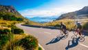 Zwei Radfahrer fahren auf einer kurvigen Bergstraße mit Blick auf eine weitläufige Bucht; die Sonne wirft lange Schatten, und die umliegende Landschaft ist von grünen Hügeln und Felsen geprägt.