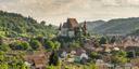 Idyllisches Dorf mit einer historischen Kirche oder Burg auf einem zentralen Hügel, umgeben von grünen Hügeln und Wäldern. Die traditionellen Gebäude mit Schrägdächern verleihen der Szenerie eine ruhige und malerische Atmosphäre.