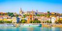 Panorama von Budapest mit Blick auf das Fischerbastei und die Matthiaskirche auf der Budaer Seite. Im Vordergrund bunte Gebäude entlang der Donau und ein Flusskreuzfahrtschiff am Ufer, dahinter historische Architektur und grüne Hügel.