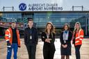 Fünf Mitarbeitende stehen vor dem Terminalgebäude des Albrecht Dürer Airport Nürnberg. Im Hintergrund ist die moderne Glasfassade des Flughafens mit dem gut sichtbaren Schriftzug „Albrecht Dürer Airport Nürnberg“ zu sehen. Die Personen tragen unterschiedliche Arbeitskleidung, darunter Business-Outfits, Uniformen und Warnschutzjacken, und repräsentieren verschiedene Tätigkeitsbereiche des Flughafens. Die Aufnahme entstand bei sonnigem Wetter im Außenbereich des Flughafens.