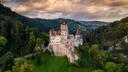 Luftaufnahme einer mittelalterlichen Burg mit roten Ziegeldächern auf einem felsigen Hügel, umgeben von dichten grünen Wäldern und sanften Berglandschaften. Über der Burg ziehen sich große, orangefarben beleuchtete Wolken über den Himmel, wodurch die Szene in warmes Abendlicht getaucht wird. Die Burg steht zentral im Bild und hebt sich deutlich von der umliegenden Natur ab.