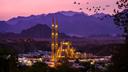 Moschee mit zwei beleuchteten Minaretten in einem Tal bei Dämmerung, umgeben von dunklen Bergsilhouetten. Der Himmel zeigt rosa und violette Farbtöne, während ein Vogelschwarm darüber hinwegfliegt.