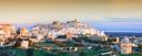 Panoramablick auf die italienische Stadt Ostuni in Apulien bei Sonnenuntergang. Die weiß getünchten Häuser schmiegen sich terrassenförmig an einen Hügel, umgeben von grünen Feldern und Gärten. Im Hintergrund erstreckt sich das tiefblaue Meer bis zum Horizont.