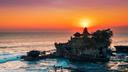 Beeindruckender Sonnenuntergang über dem Ozean mit leuchtenden Orange- und Rottönen am Himmel. Im Vordergrund befindet sich ein Felsen mit dem traditionellen balinesischen Tempel Tanah Lot, der von grüner Vegetation umgeben ist. Der Tempel hat mehrere gestufte Dächer. Unten schlagen Wellen gegen die Felsen, während Menschen rund um den Fuß des Felsens spazieren und die Aussicht genießen.