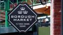 Schild des Borough Market mit weißer Schrift auf schwarzem Hintergrund und drei Kronensymbolen darüber. Im Hintergrund sind grüne Metallstrukturen und eine Backsteinwand zu sehen, die auf eine Außen- oder halbüberdachte Umgebung hinweisen.