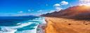 Panoramablick auf einen Strand mit goldenem Sand, sanften Wellen und Bergen im Hintergrund unter einem blauen Himmel mit vereinzelten Wolken.
