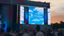 Besucherinnen und Besucher stehen vor einer großen Festivalbühne auf dem Gelände des Albrecht Dürer Airport Nürnberg. Auf den LED-Bildschirmen wird das Logo des Airports vor einem Himmelmotiv angezeigt. Die Aufnahme entstand während eines Festivals bei Sonnenuntergang, der Himmel ist in warme Orange- und Blautöne getaucht. Vor der Bühne versammelt sich eine große Menschenmenge, während die Festivalatmosphäre durch die beleuchtete Bühne und die offene Flughafenkulisse geprägt wird. Auf dem Bühnendach ist der Schriftzug „Festival Summer“ zu sehen.