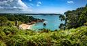 Küstenlandschaft auf der Kanalinsel Jersey mit türkisblauem Meer, einer kleinen Sandbucht, felsigen Klippen und üppiger grüner Vegetation unter einem teilweise bewölkten Himmel.