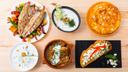 Verschiedene traditionelle bulgarische Gerichte auf einem Holztisch darunter gegrillter Fisch mit Gemüse eine Joghurtsuppe ein Eintopf im Tontopf sowie Gebäck und belegtes Brot mit Weißkäse.