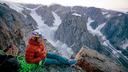 Bergsteiger sitzt auf einem Felsvorsprung inmitten einer dramatischen Gebirgslandschaft mit Gletschern und steilen Klippen. Er trägt eine rote Jacke, blaue Hose und Kletterausrüstung. Im Hintergrund erstreckt sich eine weite, schneebedeckte Landschaft unter einem sanften Himmel.