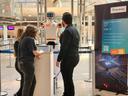 Zwei Personen stehen vor einem kleinen humanoiden Roboter und interagieren mit einem Display auf einem Podest in einer modernen, lichtdurchfluteten Innenhalle. Rechts daneben befindet sich ein Aufsteller mit Fraunhofer-Logo und Informationen zu „Edge AI“ und maschinellem Lernen für eingebettete Systeme.