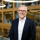 Ein Mann steht in einem modernen Bürogebäude mit viel Holz und Glas. Er trägt einen dunklen Anzug und ein weißes Hemd. Hinter ihm sind offen gestaltete Arbeitsbereiche, rechteckige Fensterrahmen aus Holz sowie große, ringförmige Deckenleuchten zu sehen.