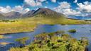 Landschaftsaufnahme einer grünen Insel, umgeben von tiefblauem Wasser. Im Hintergrund erstrecken sich sanfte Hügel und Berge unter einem teils bewölkten Himmel. Die üppige Vegetation der Insel steht im Kontrast zum Wasser und vermittelt eine ruhige, naturnahe Atmosphäre.