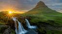 Kirkjufell-Berg in Island bei Sonnenuntergang. Die Sonne strahlt hinter dem Berg hervor, während im Vordergrund ein Wasserfall in einen Fluss fließt, umgeben von saftig grünem Gras.