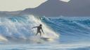 Surfer reitet eine große Welle im Ozean, umgeben von spritzendem Wasser. Im Hintergrund sind Berge und ein klarer Himmel zu sehen.