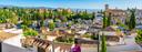 Panorama des Albayzín-Viertels in Granada, Spanien, mit weißen Häusern und Terrakottadächern im Vordergrund. Im Hintergrund thront die Alhambra auf einem bewaldeten Hügel, dahinter die Sierra Nevada unter klarem Himmel.