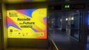Beleuchtete Werbefläche im Innenbereich des Albrecht Dürer Airport Nürnberg mit einer Anzeige für das Nürnberg Digital Festival. Das farbenfrohe Motiv in Gelb, Blau, Pink und Orange trägt den Slogan „Recode the Future“ sowie den Veranstaltungszeitraum „22.6.–2.7.2026“. Im unteren Bereich sind verschiedene Partnerlogos und der Hinweis „Scan & Discover More Than 200 Events“ mit QR-Code abgebildet. Die Werbefläche befindet sich in einem Terminalgang in der Nähe eines Check-in-Bereichs und ist von den gedämpft beleuchteten Flughafenräumlichkeiten umgeben.