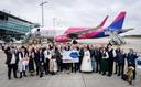 Menschen stehen vor einem Wizz Air Flugzeug auf dem Rollfeld, einige in traditioneller Kleidung, andere mit einem großen Schild – im Hintergrund Flughafeninfrastruktur.