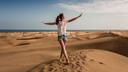 Person mit ausgestreckten Armen steht auf einer Sanddüne in einer weiten Wüstenlandschaft. Im Hintergrund erstrecken sich Dünen bis zum Horizont, wo Meer und Himmel aufeinandertreffen.