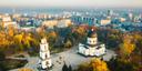 Luftaufnahme einer Stadt mit einer markanten Kirche mit großer Kuppel und Säulen sowie einem mehrstöckigen weißen Glockenturm mit schwarzer Kuppel. Umgeben von herbstlich gefärbten Bäumen, Wegen und Wohngebäuden im Hintergrund.
