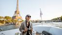 Person mit Sonnenbrille und Kamera sitzt auf einem Boot auf der Seine in Paris. Im Hintergrund erhebt sich der Eiffelturm, während eine Brücke den Fluss überspannt. Die Szene ist von warmem Tageslicht geprägt und zeigt die französische Hauptstadt aus der Perspektive einer Bootsfahrt.