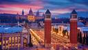 Abendaufnahme von Barcelona mit den beiden venezianischen Türmen an der Plaça d’Espanya, einer hell erleuchteten Allee und dem beleuchteten Palau Nacional vor einem farbenprächtigen Himmel.