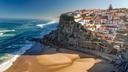 Küstenort in Portugal mit weißen Häusern und roten Dächern, die sich entlang einer felsigen Klippe erstrecken. Darunter liegt ein Sandstrand, an dem türkisfarbenes Meerwasser anlandet. Der Himmel ist klar und blau.