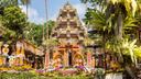 Traditioneller balinesischer Tempel mit kunstvollen Schnitzereien und lebhaften Dekorationen, umgeben von üppigem Grün. Vor dem Tempel befinden sich zahlreiche bunte Blumenarrangements, die eine festliche Atmosphäre schaffen. Der Himmel ist klar mit wenigen Wolken im Hintergrund.