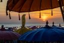 Strandszene bei Sonnenuntergang mit bunten Sonnenschirmen im Vordergrund; die untergehende Sonne taucht Himmel und Meer in warmes Orange, während Menschen am Strand und im Wasser entspannen.