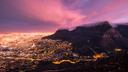 Die Stadt erstreckt sich bei Nacht in einem Meer aus goldenen Lichtern. Rechts ragt ein großer, flachgedeckter Berg empor, dessen Gipfel teilweise von Nebel bedeckt ist. Der Abendhimmel leuchtet darüber in leuchtendem Rosa und Lila. Zahlreiche Straßen und Wohnviertel sind als helle Lichtpunkte klar zu erkennen.