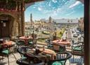 Outdoor-Café mit bunten, gemusterten Tischdecken, Blick auf einen belebten Stadtplatz mit moderner und historischer Architektur, darunter eine Moschee mit Minarett; im Hintergrund Gebäude, Autos und ein teils bewölkter Himmel.