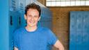 Ein junger Mann mit lockigen Haaren und einem blauen T-Shirt lächelt fröhlich in die Kamera. Er steht locker neben einer Reihe blauer Spinde. Im Hintergrund erkennt man eine Ziegelwand und ein helles Fenster.