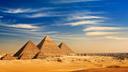 Das Bild zeigt die Pyramiden von Gizeh in Ägypten. Im Vordergrund sind drei große Pyramiden zu sehen, die sich majestätisch aus der Wüste erheben. Der Himmel ist klar und blau mit einigen dünnen, weißen Wolkenstreifen. Im Hintergrund ist eine moderne Stadtlandschaft zu erkennen, die einen Kontrast zur antiken Architektur der Pyramiden bildet.