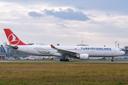 Ein Airbus A330 von Turkish Airlines rollt auf der Start- oder Landebahn des Albrecht Dürer Airport Nürnberg. Das Flugzeug ist seitlich zu sehen, mit rotem Leitwerk und dem weißen Logo der Airline. Im Hintergrund stehen weitere Flugzeuge und Gebäude des Flughafens unter bewölktem Himmel.
