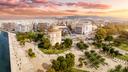 Panoramablick auf Thessaloniki bei Sonnenuntergang mit dem Weißen Turm im Vordergrund, umgeben von Grünanlagen und Promenade am Meer, dahinter dichte Stadtbebauung bis zu den Hügeln.
