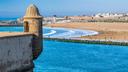 Blick von einer historischen Festungsmauer auf eine weite Atlantikbucht bei Rabat. Im Vordergrund steht ein steinerner Wachturm mit kuppelförmigem Dach. Dahinter erstrecken sich türkisblaues Wasser, ein breiter Sandstrand mit Spaziergängern und sanften Wellen sowie die Stadtmauer und Bebauung entlang der Küste unter klarem blauem Himmel.