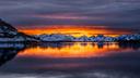 Ein ruhiger See spiegelt eine schneebedeckte Bergkette wider. Am Horizont leuchtet ein goldener Sonnenuntergang unter dunklen Wolken. Links fällt ein felsiger, schneebedeckter Hang zum Wasser ab. Die Wasseroberfläche zeigt klar die Farben des Himmels und der Berge.