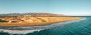 Panoramablick auf eine Küstenlandschaft mit Sanddünen entlang der Uferlinie, sanften Wellen, Bergen im Hintergrund und einer kleinen Siedlung am Meer.