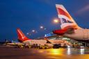 Mehrere Flugzeuge stehen auf einem beleuchteten Flughafen bei Dämmerung. Im Vordergrund ist das Heck eines British Airways Flugzeugs zu sehen, im Hintergrund ist eine Corendon Airlines Maschine und ein Ryanair Flugzeug geparkt. Bodenfahrzeuge bewegen sich um die Flugzeuge herum, der Himmel ist dunkelblau.