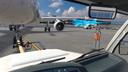 An der Wand eines Raums montierter Bildschirm mit einer Simulation oder VR-Darstellung eines Flugzeugs auf dem Vorfeld. Auf dem Bildschirm sind das Bugfahrwerk und die Triebwerke eines Flugzeugs aus der Bodenabfertigungsperspektive zu sehen. Links neben dem Bildschirm hängt eine orangefarbene Warnjacke, darüber befinden sich gerahmte Informations- und Lagepläne.