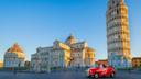 Der Schiefe Turm von Pisa zusammen mit dem Dom und dem Baptisterium. Davor steht ein alter roter Wagen mit der Aufschrift „Luigi“ auf einer leeren Straße.