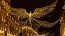 Nächtliche Stadtstraße mit großen, goldenen Engelsfiguren aus Lichterketten, deren ausgebreitete Flügel über die Straße reichen. Die umliegenden Gebäude sind ebenfalls beleuchtet und verstärken die festliche Stimmung.