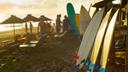 Strandszene bei Sonnenuntergang mit mehreren Surfboards, die an einer Holzstruktur auf der rechten Seite lehnen. Im Hintergrund sitzen und stehen Menschen unter großen Strandsonnenschirmen auf Liegestühlen. Das Meer mit sanften Wellen und ein wolkiger Himmel, der vom Sonnenuntergang beleuchtet wird, sind in der Ferne sichtbar.