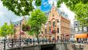 Historische Grachtenlandschaft in Amsterdam mit einer Backsteinbrücke über einen Kanal und zahlreichen abgestellten Fahrrädern entlang des Geländers. Im Hintergrund steht ein repräsentatives Gebäude aus rotem Backstein mit weißen Verzierungen und giebelständiger Fassade. Blühende Blumenampeln, große grüne Bäume und der blaue Himmel verleihen der Szene eine typisch niederländische und lebendige Atmosphäre.
