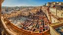 Panoramablick über die historische Gerberei von Fès mit zahlreichen runden und rechteckigen Steinbecken, die mit unterschiedlich gefärbten Flüssigkeiten gefüllt sind. Umgeben von dicht bebauten Sandsteinhäusern der Medina arbeiten einzelne Personen zwischen den Becken. Die Szene wird von warmem Sonnenlicht beleuchtet und vermittelt einen Eindruck des traditionellen Handwerks.