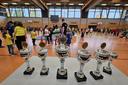 Sechs verschieden große silbern-glänzende Pokal stehen auf einem Tisch in der Turnhalle. Im Hintergrund machen Kinder Sport.